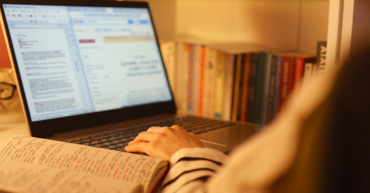 A person studying online with a laptop and open book in a cozy home setting, surrounded by shelves of books.
