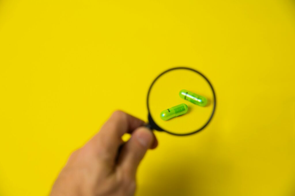 From above of crop unrecognizable male holding magnifying glass over green pills on yellow background in studio